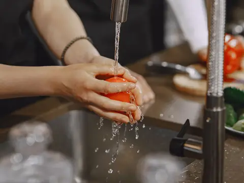A tomato rinsed under running tap water at a kitchen sink